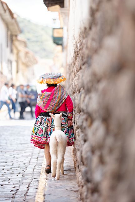 Quechua woman in colorful attire with a llama on a street in Cusco, Peru, showcasing traditional culture.