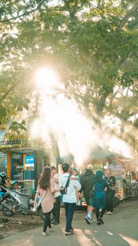 Street scene in Pontianak, Indonesia with sunlight streaming through trees and people walking.