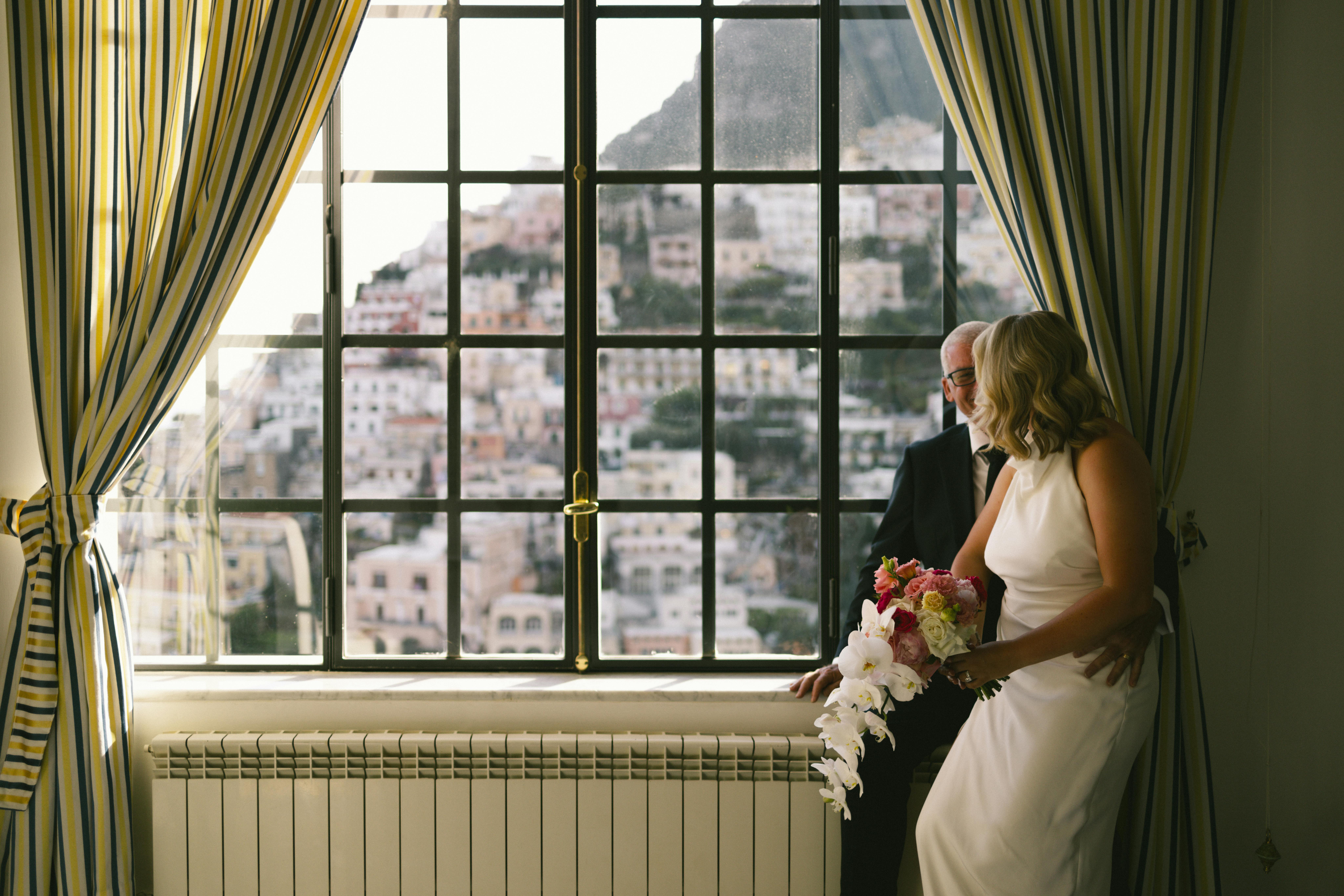 A bride and groom in elegant attire with a picturesque view of Positano, Italy through the window.