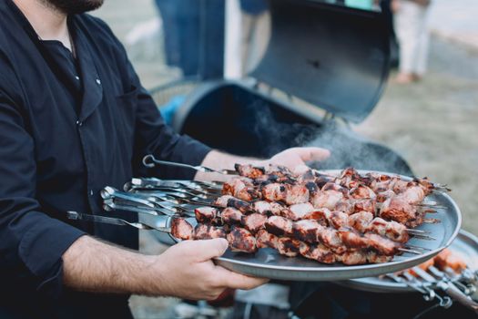 A man holding a tray of freshly grilled meat skewers with smoke rising outdoors.