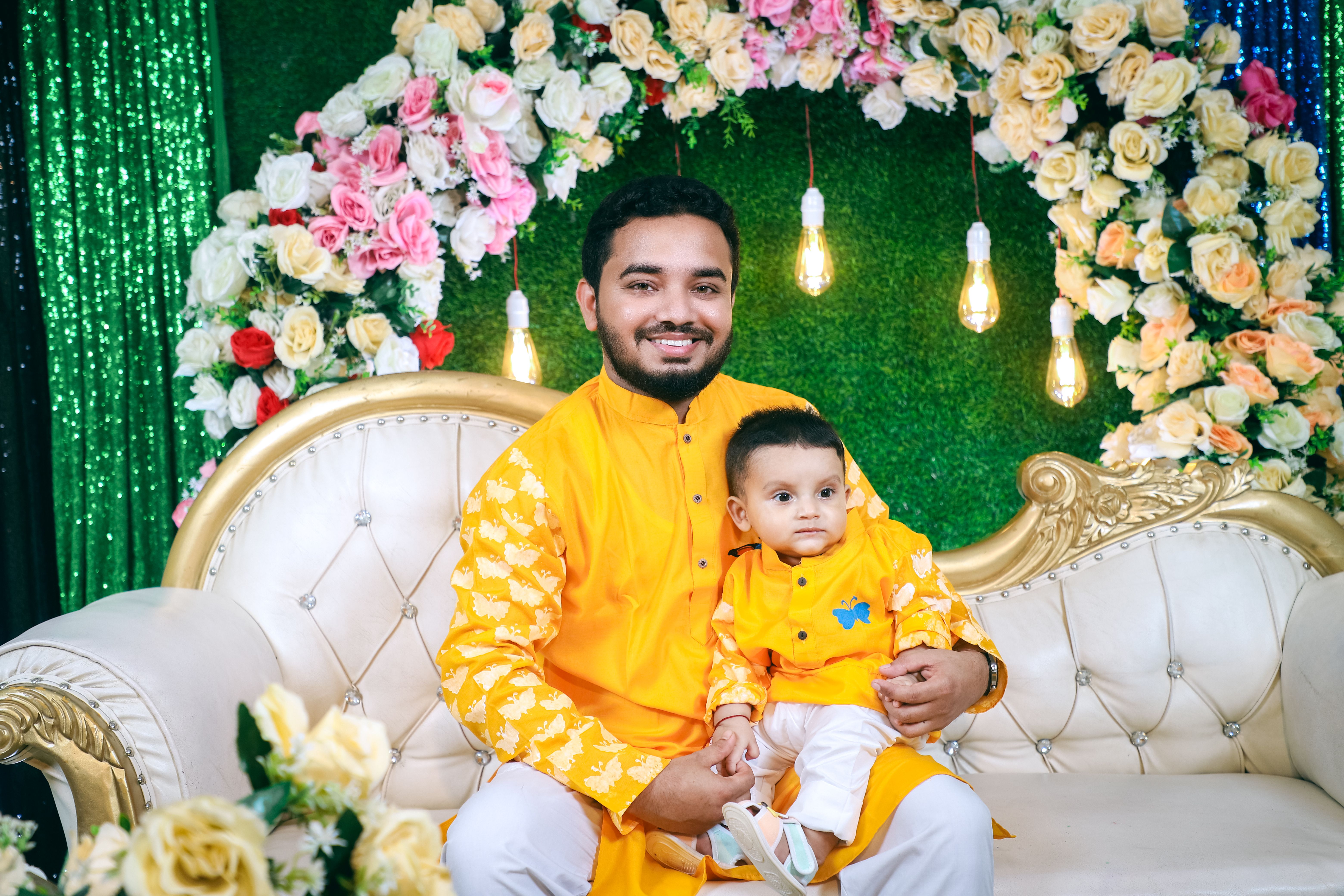 A father and son wearing bright traditional outfits sitting on a decorated sofa enjoying family time.