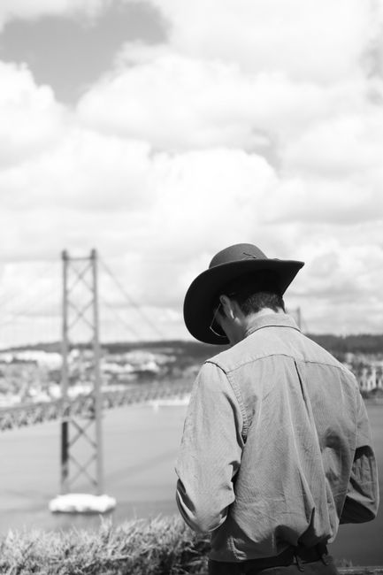 Black and white photo of a man in Lisbon, viewing the 25 de Abril Bridge with a cloudy sky.