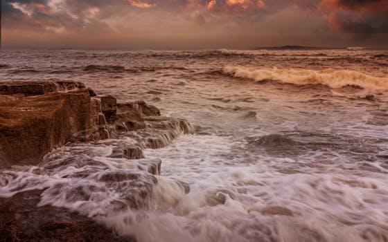 Waves crash against rocky coast under a colorful sunset sky, creating a dramatic seascape.
