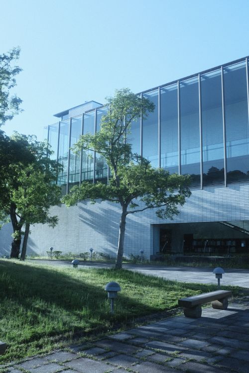 Free Exterior view of a modern glass building with a bench surrounded by trees in daylight. Stock Photo