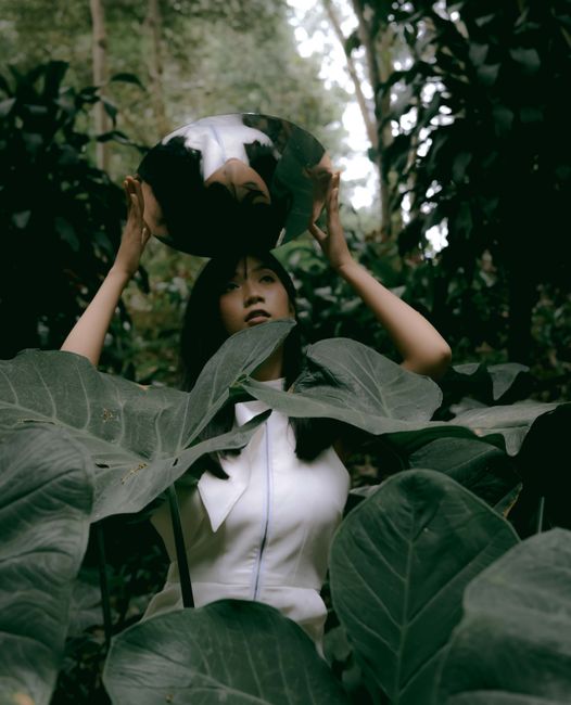 A woman in a jungle poses with a reflective mirror amidst lush greenery.