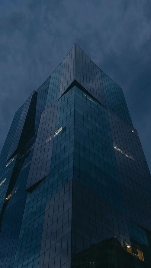 Free A towering modern skyscraper with glass facade under an overcast night sky. Stock Photo