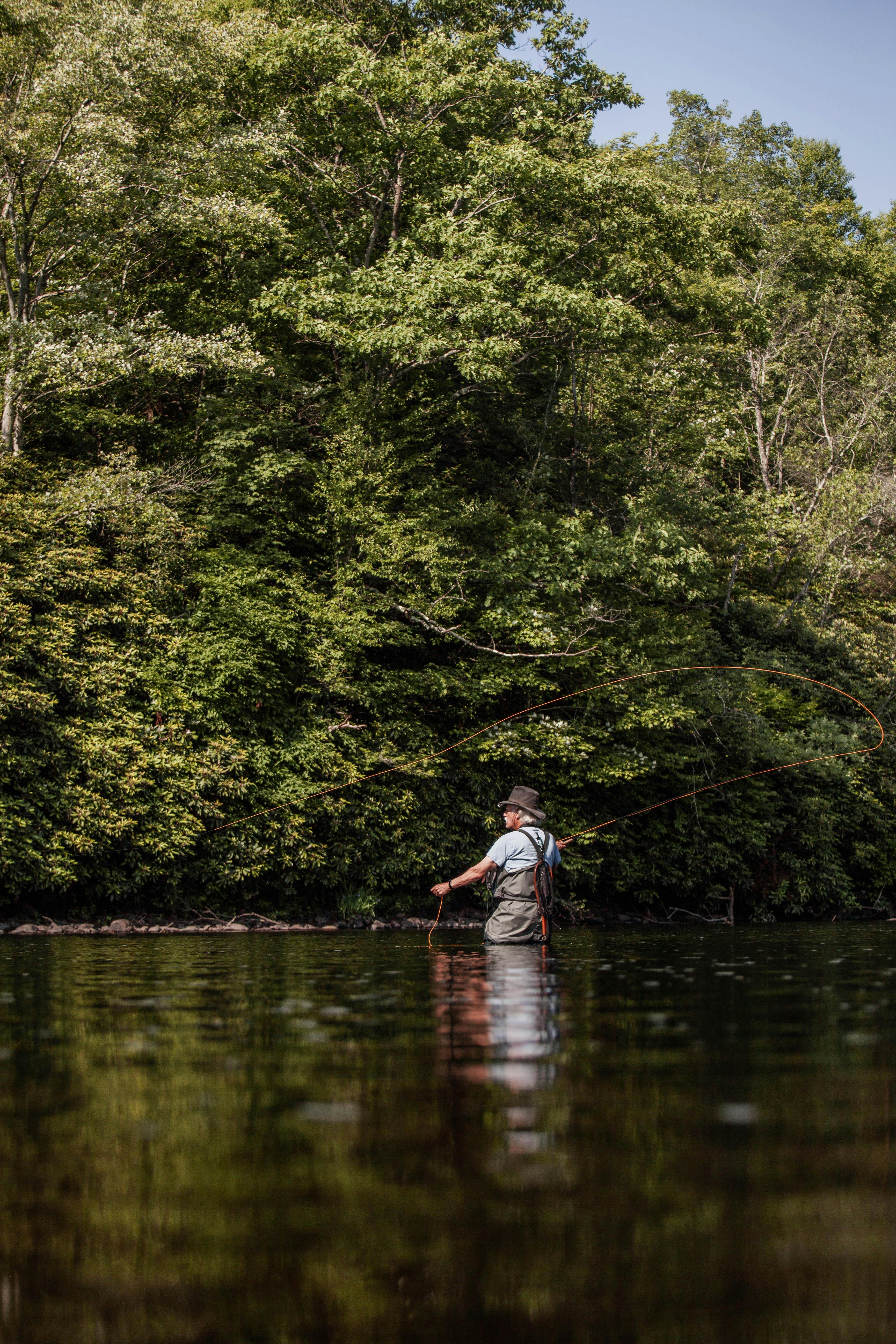 An adult man enjoys fly fishing in a river surrounded by lush green forest in summer.