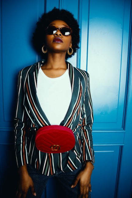 Fashionable woman posing with confidence in a striped blazer and red waist bag against a blue wall.