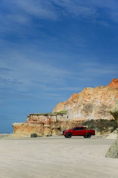 A red pick-up truck parked on a picturesque beach with colorful cliffs under a clear blue sky.