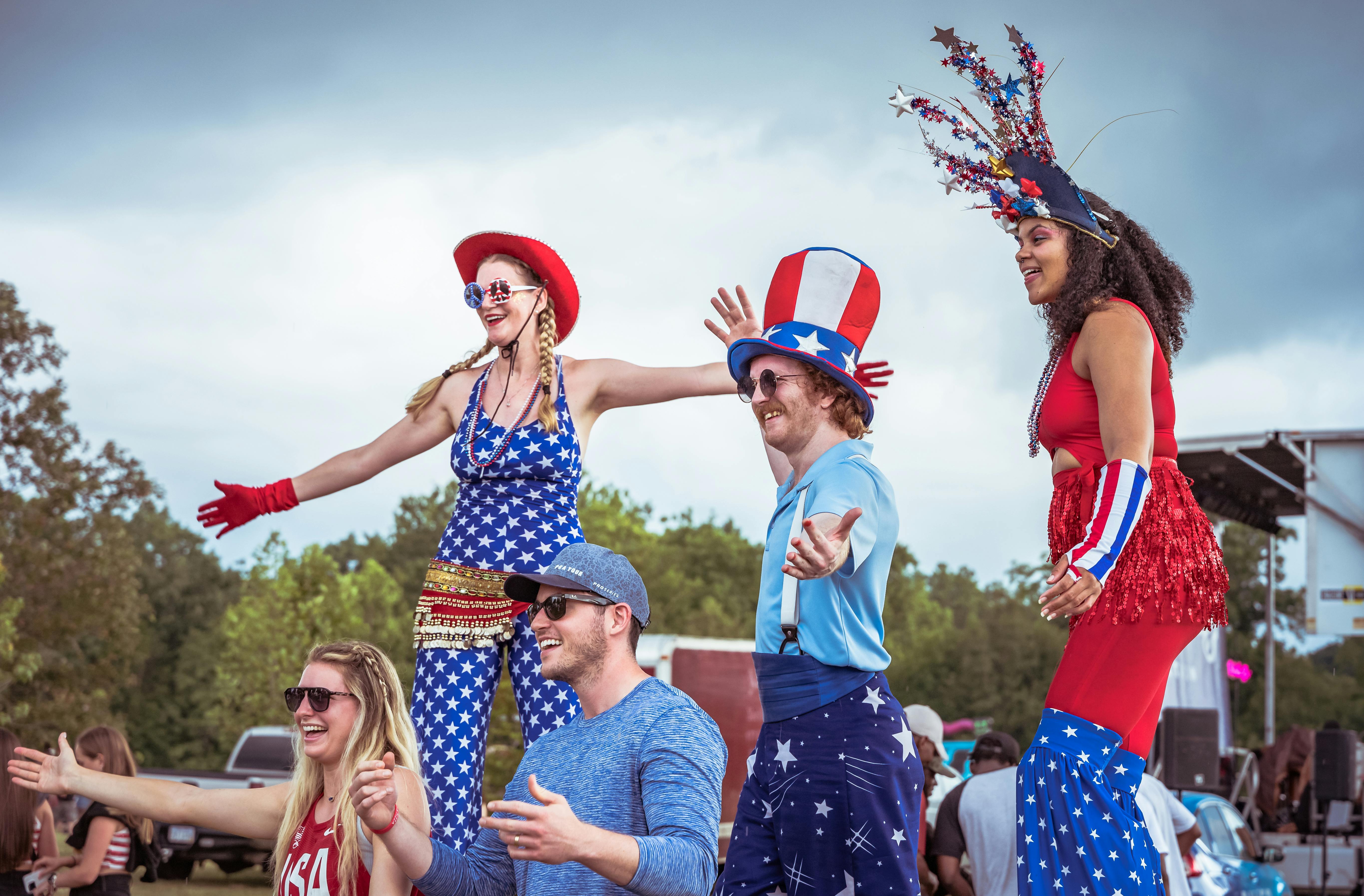 Colorful outdoor event featuring performers on stilts in patriotic attire, smiling and engaging with a cheerful crowd.