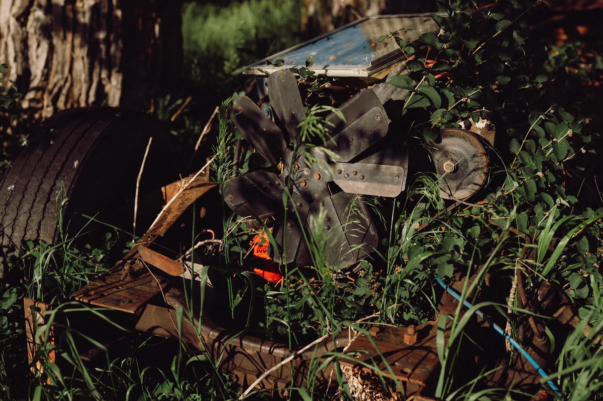 An old, rusty machine with propellers sits abandoned and overgrown by shrubs and grass.