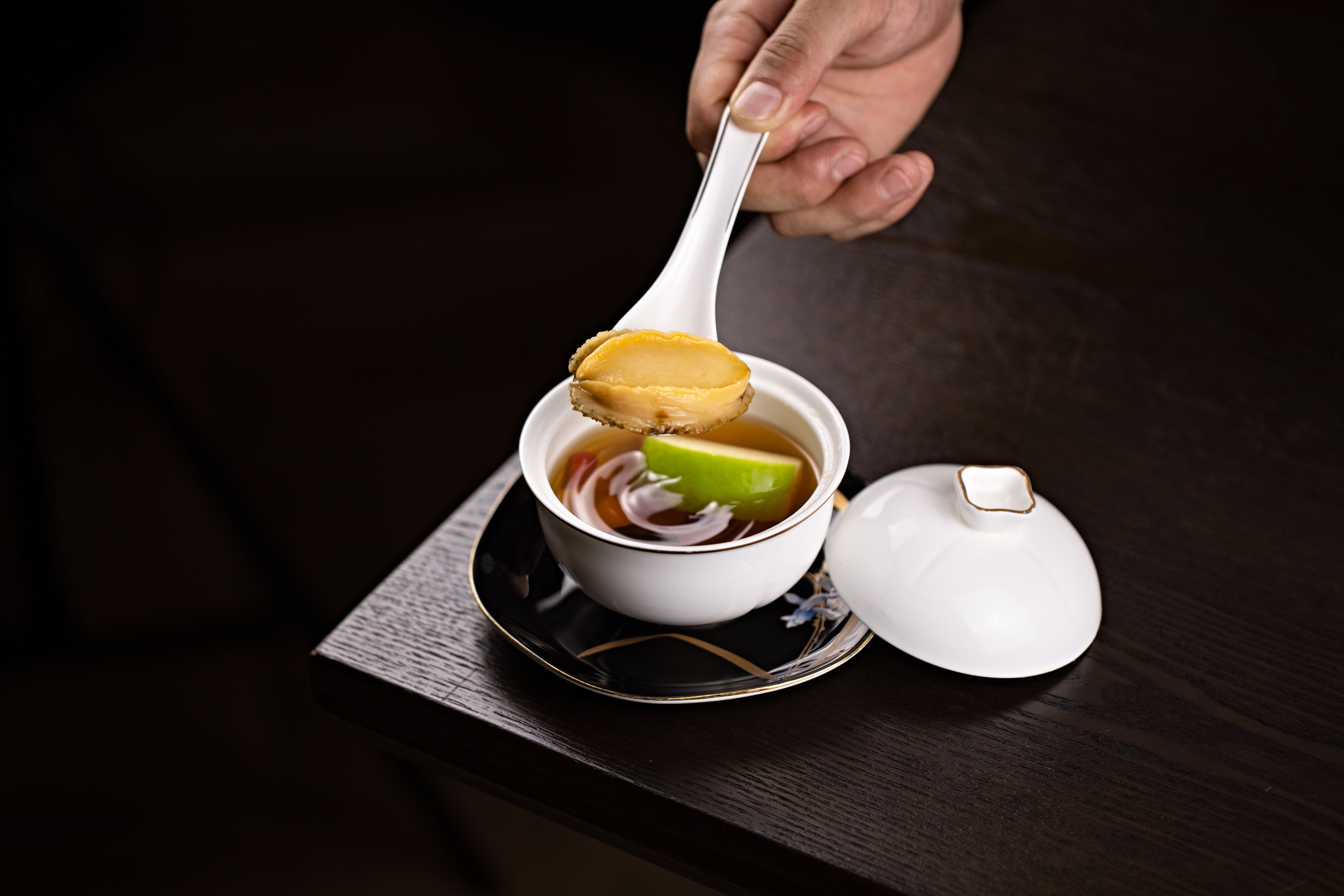 A gourmet dish featuring abalone soup, elegantly served in a white porcelain cup with a spoon.