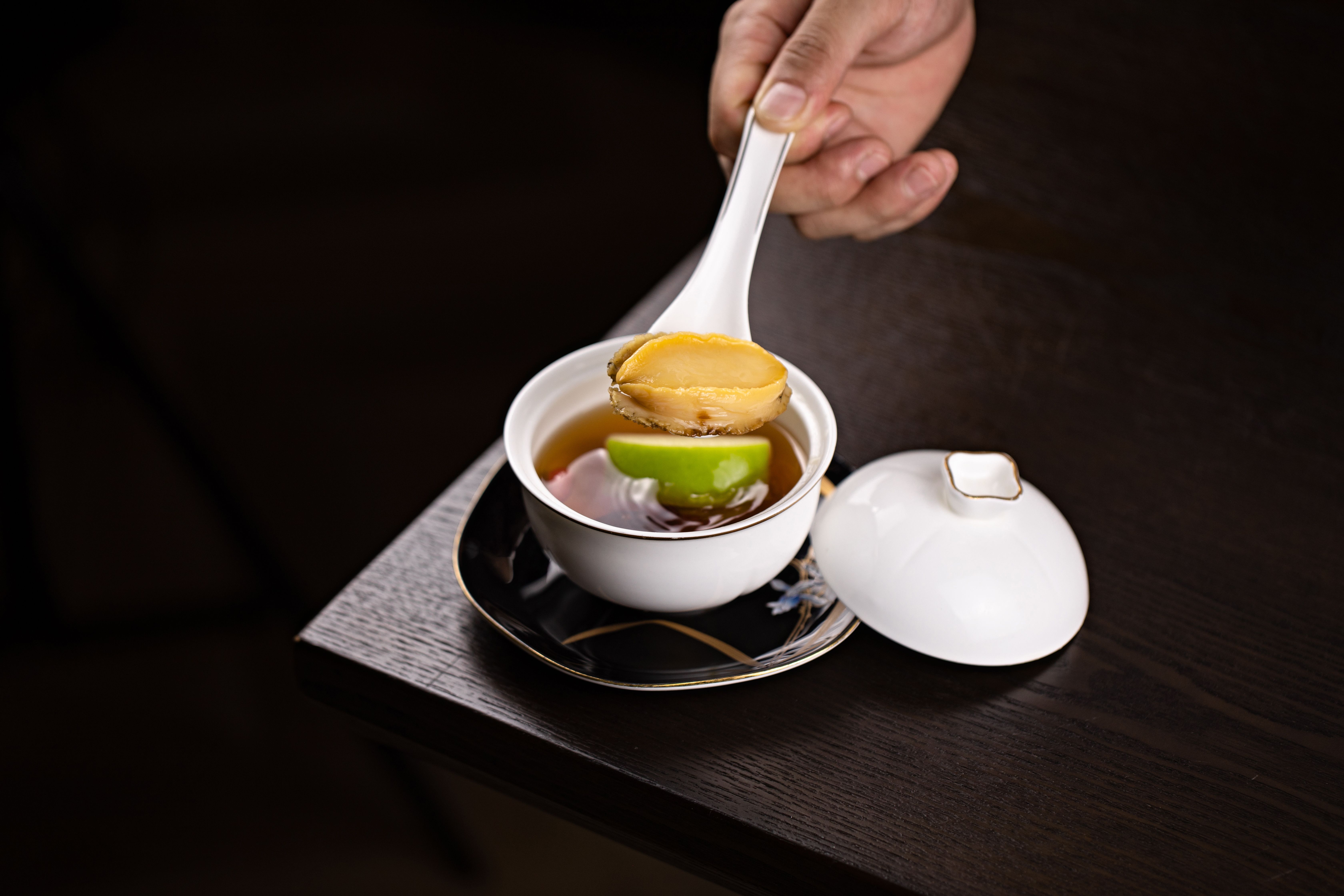 A sophisticated presentation of soup with abalone, carefully plated on dark wood.