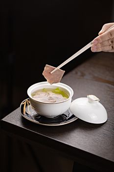 A close-up of a gourmet Asian soup with chopsticks picking up tender meat in an elegant setting.