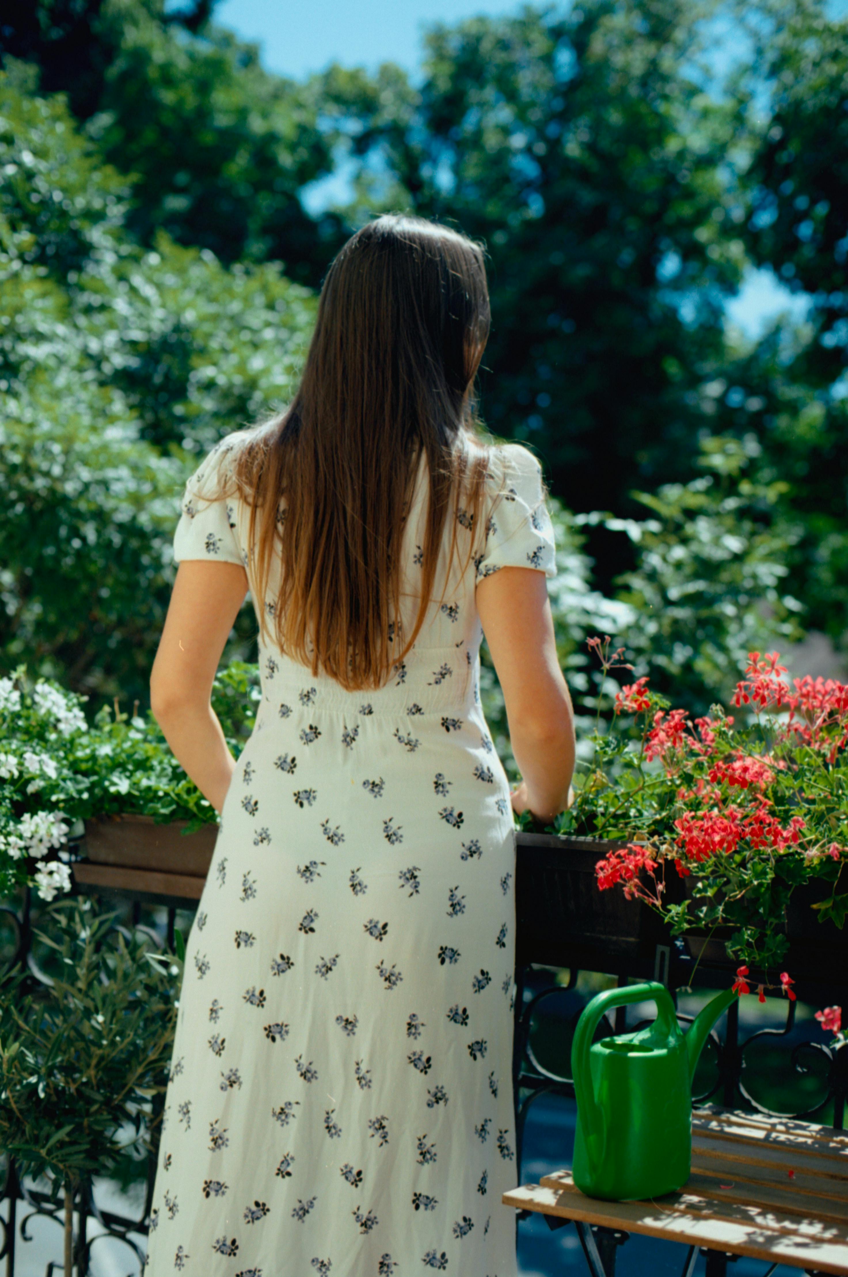 A woman in a white floral dress stands on a vibrant, flower-filled balcony on a sunny day.