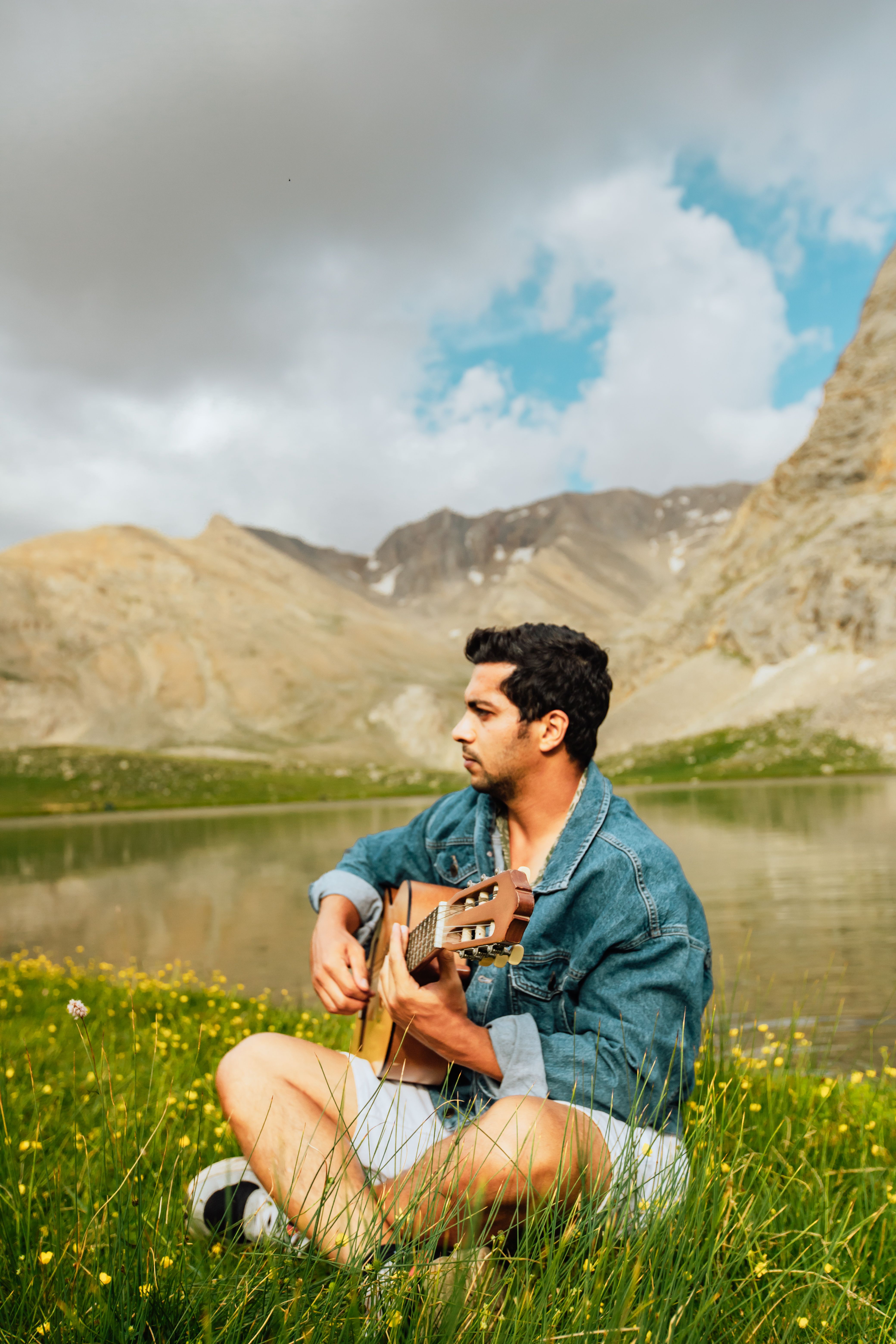 Young man in denim jacket plays acoustic guitar by a serene mountain lake in Madenköy, Türkiye
