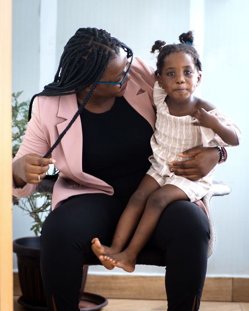 Free A mother wearing a stylish suit jacket holds her daughter on her lap indoors, showcasing a charming moment. Stock Photo