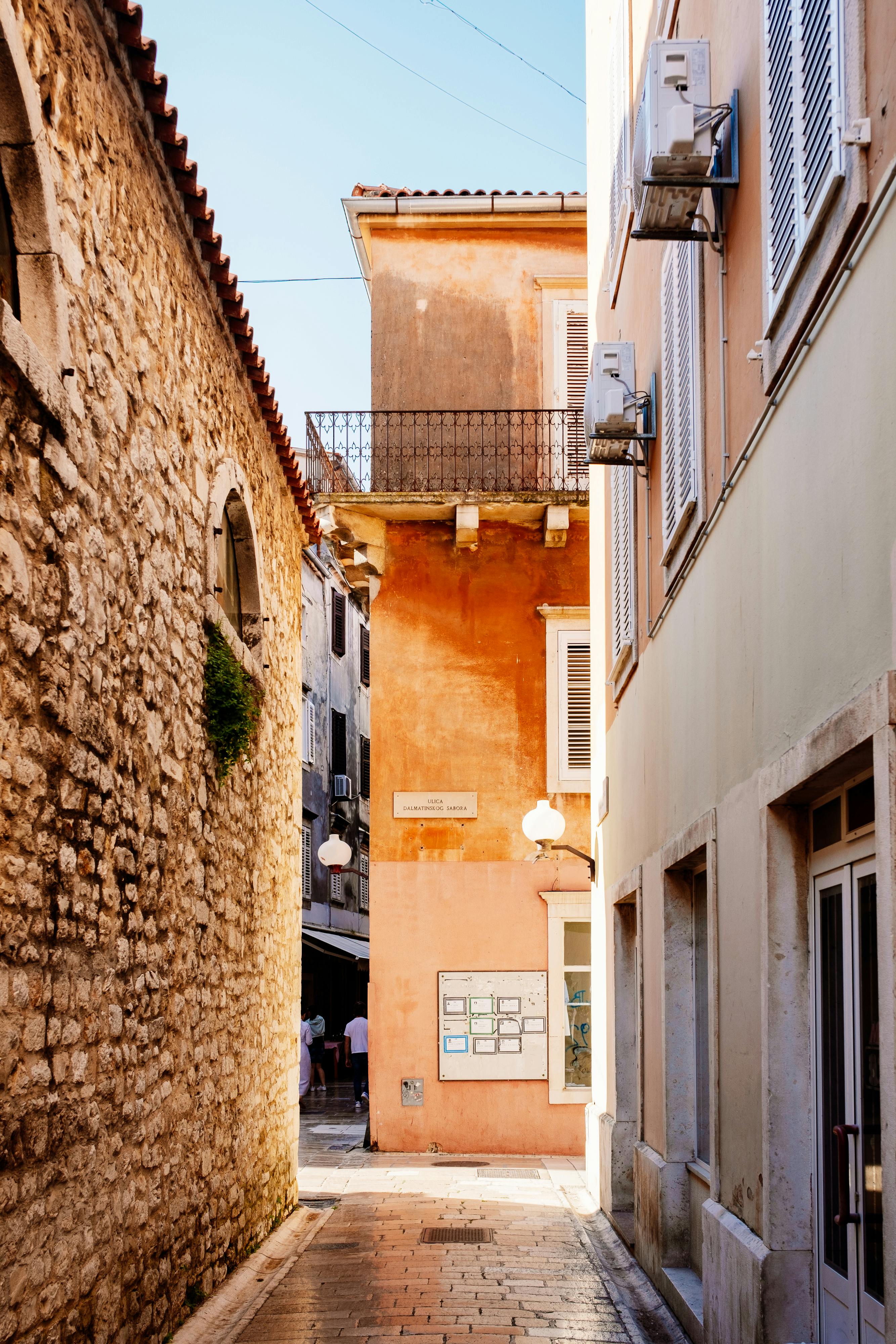 Sunlit narrow alley between stone and plaster buildings in a Mediterranean town.