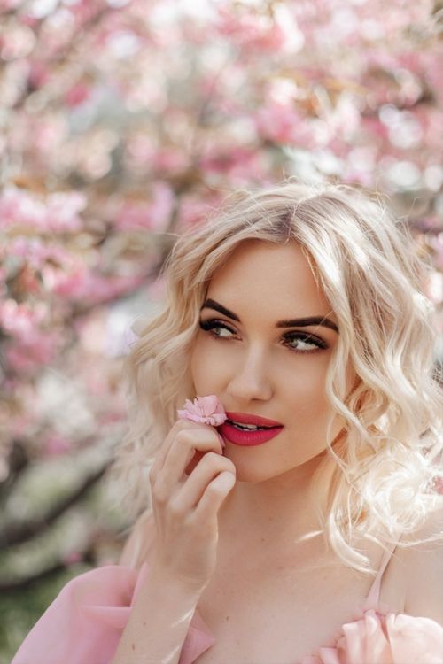 Free Portrait of a young blonde woman in a pink dress, surrounded by spring blossoms in a park. Stock Photo
