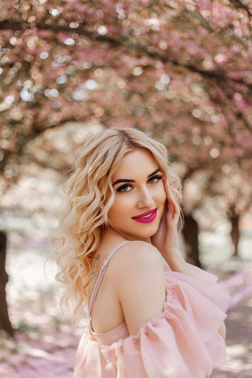 Free Elegant young woman posing in a pink dress under cherry blossoms with a bright smile. Stock Photo
