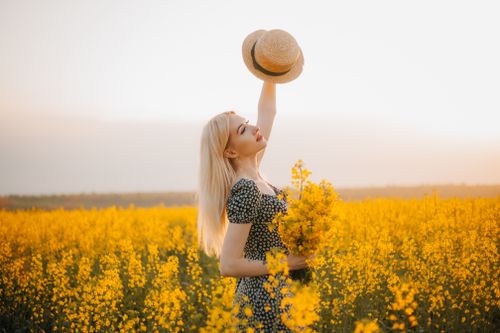 Free A woman holding a hat and flowers stands joyfully in a vibrant field during sunset. Stock Photo