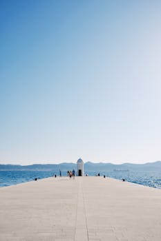 People walking on a pier towards a lighthouse with a clear blue sky.