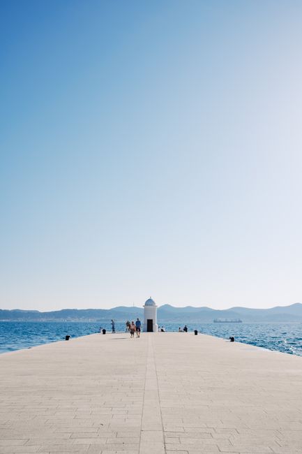 People walking on a pier towards a lighthouse with a clear blue sky.