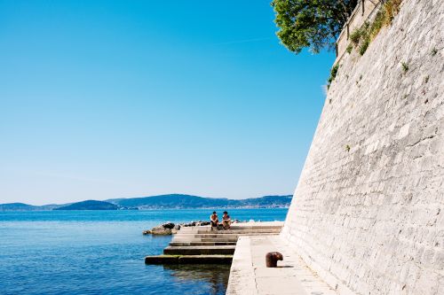 Free A sunny day with people sitting by the seawall in Zadar, Croatia, with a clear sky and sea view. Stock Photo
