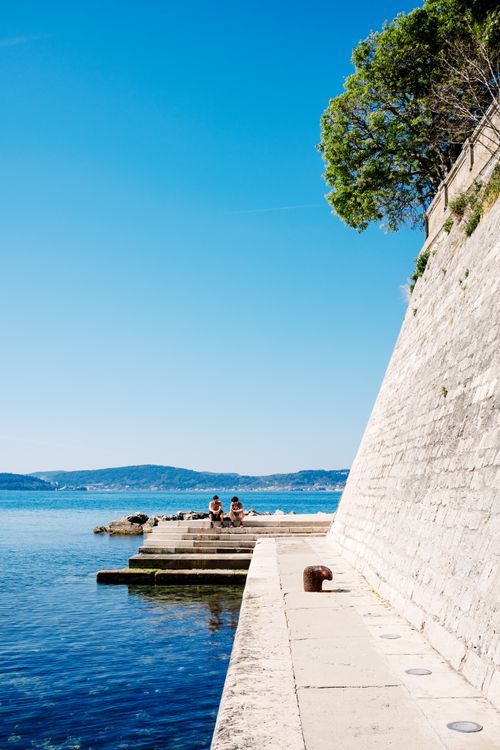 Free A peaceful promenade by the sea featuring a stone wall and clear blue waters. Stock Photo