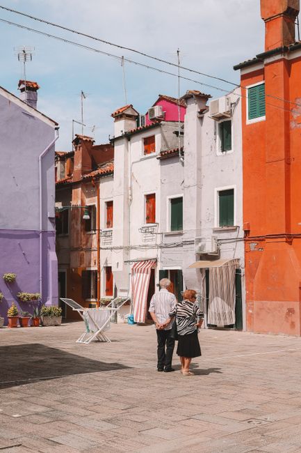 An elderly couple strolls through a vibrant square in Venice, Italy, surrounded by colorful townhouses.