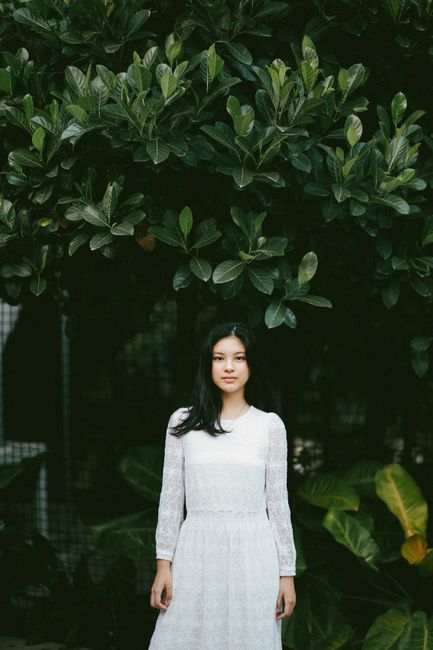 Portrait of a woman in a white dress standing against a backdrop of lush green foliage.