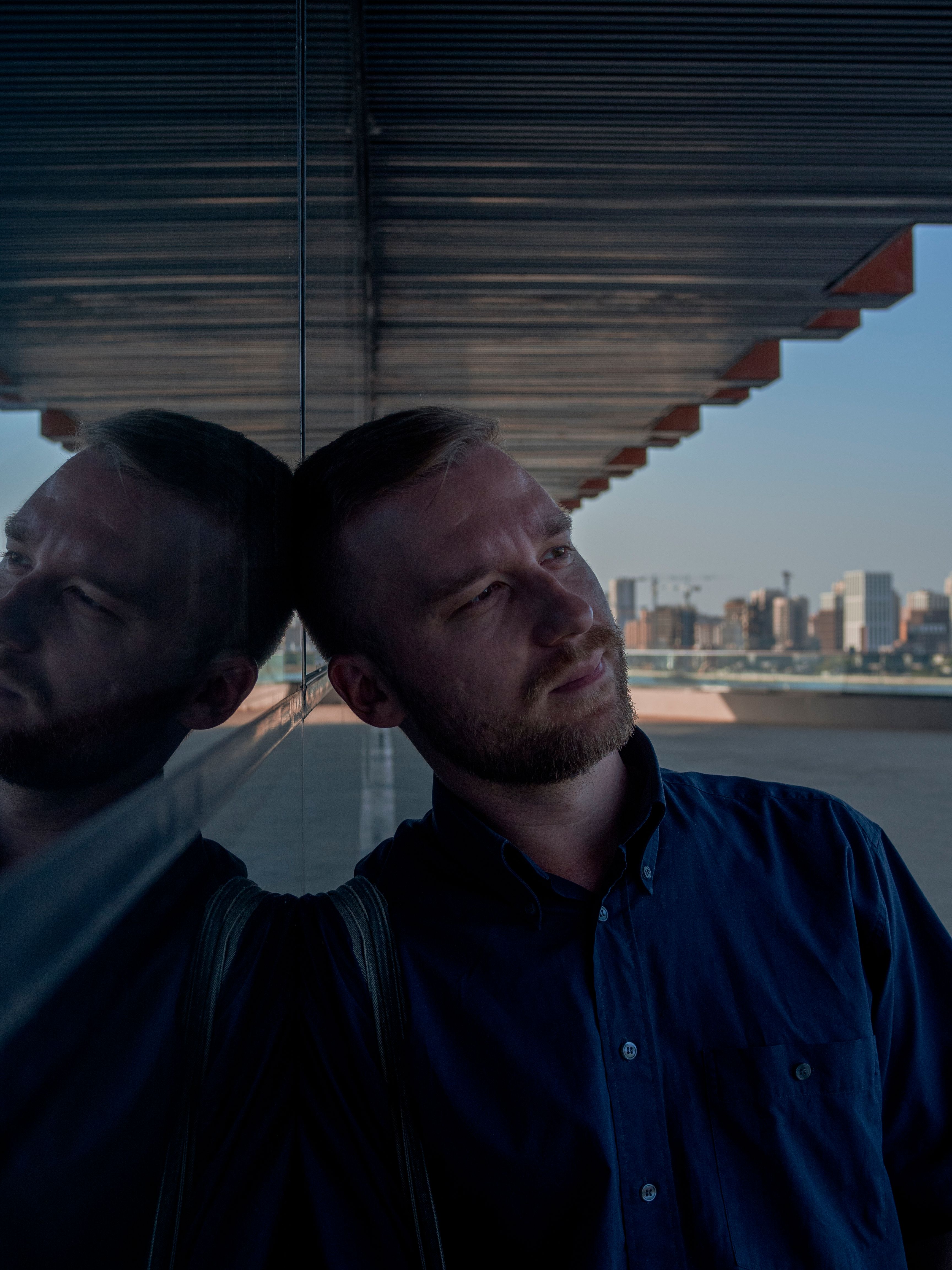 Portrait of a man leaning on a reflective wall with a city skyline in the background.