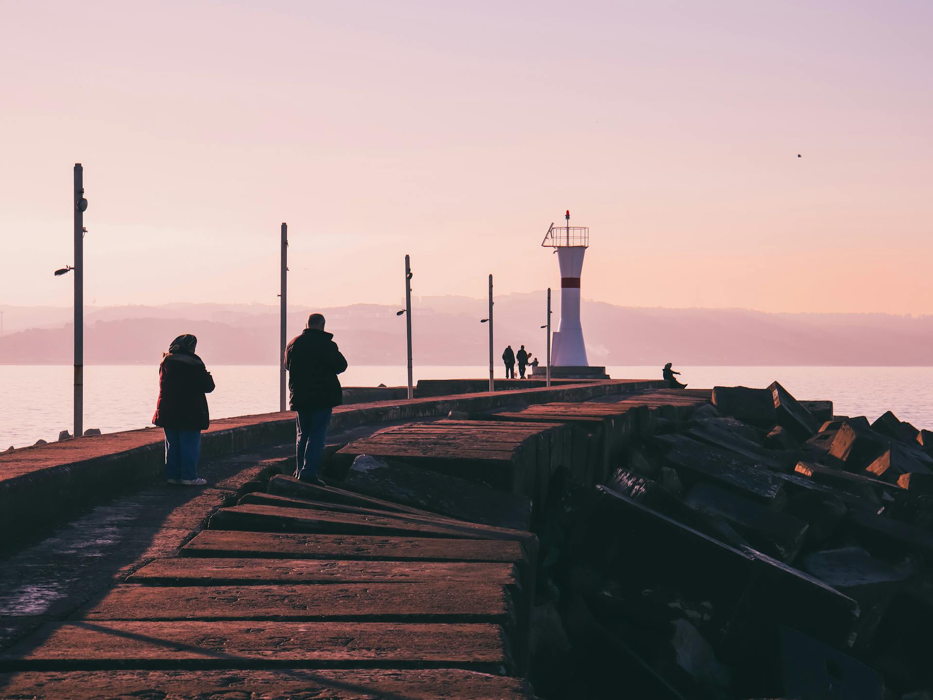 People walking along a pier near a lighthouse at sunrise by the ocean.