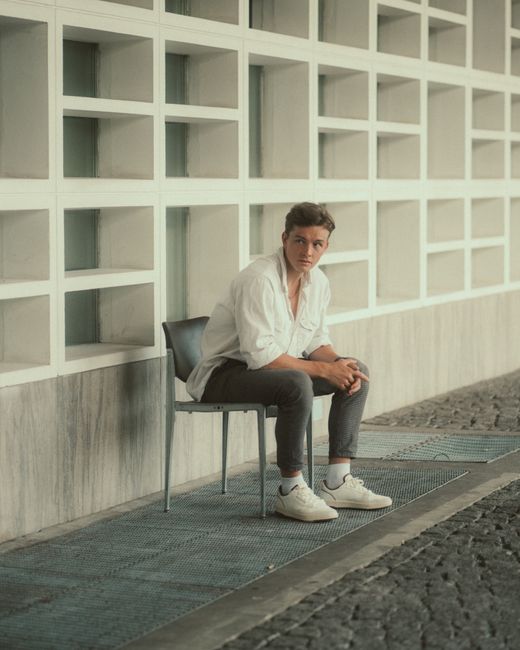A young man poses against modern architecture in Berlin, showcasing urban fashion with a casual style.