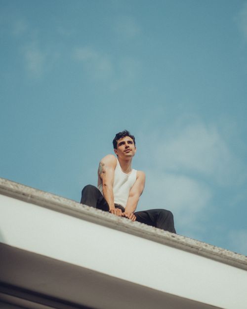 Free A man in a tank top sits on a rooftop against a clear blue sky in Berlin, capturing urban exploration. Stock Photo