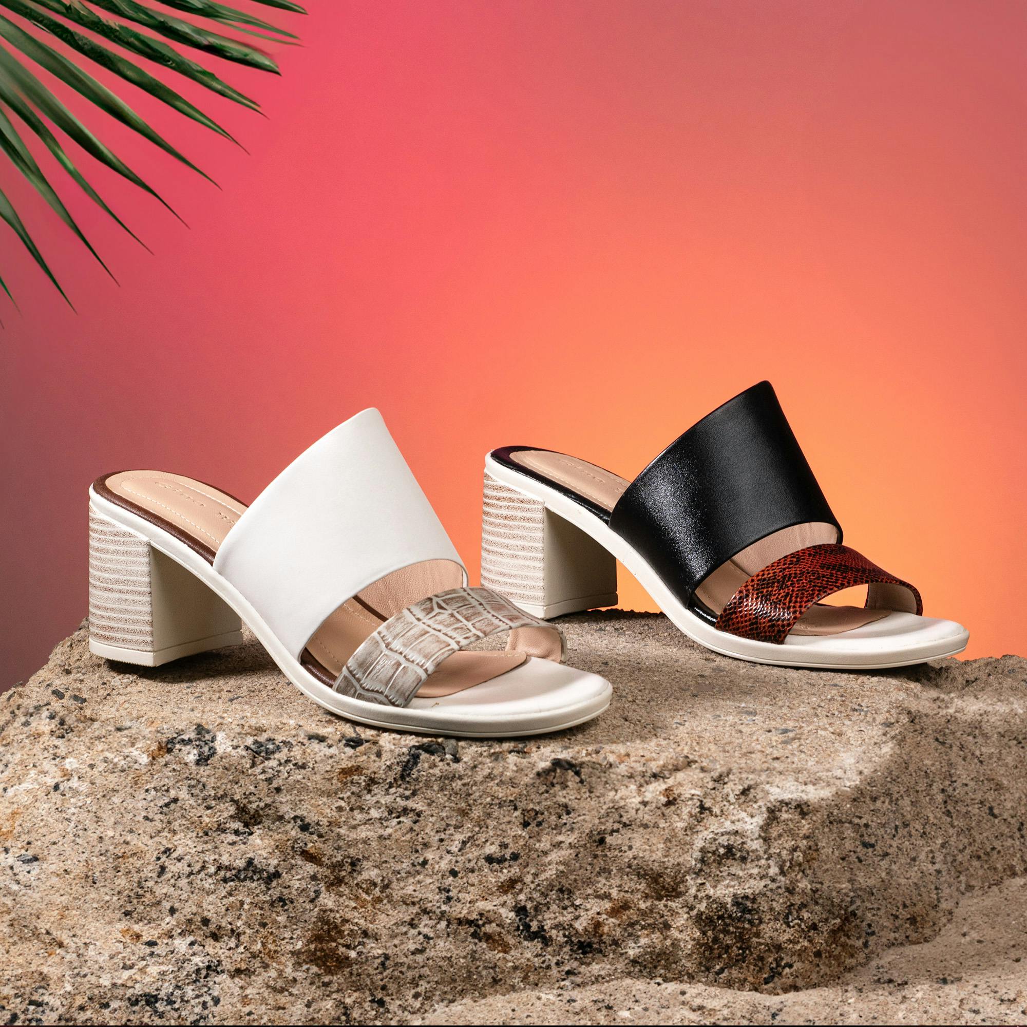 Elegant black and white block-heeled sandals displayed on rock against gradient backdrop.