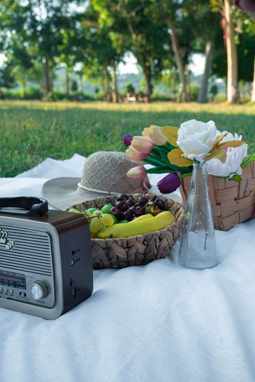 Free A serene outdoor picnic setup featuring fruits, flowers, radio, and sun hat on a blanket. Stock Photo