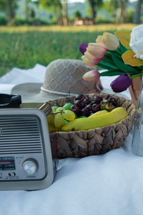 Free Charming picnic setup with a vintage radio, fruit basket, and a sun hat in a lush park setting. Stock Photo