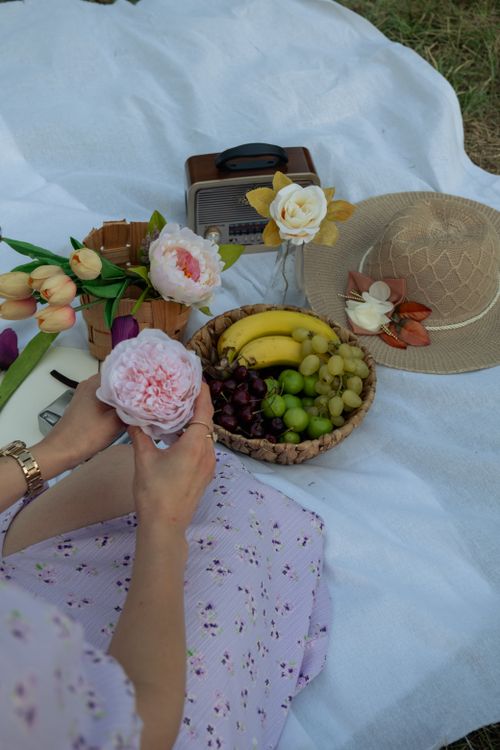 Free A serene picnic scene with fruits, flowers, and a sun hat on a white blanket. Stock Photo