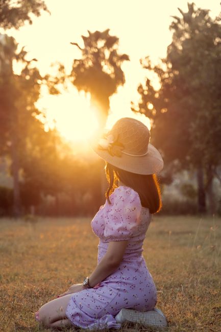 A woman in a floral dress and sunhat enjoys a peaceful sunset in a park, creating a serene moment.