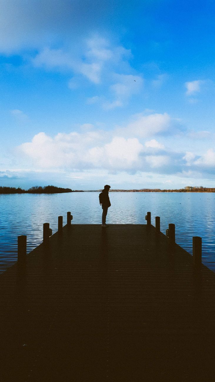 Silhouette of a man standing on a wooden pier over a calm lake under a blue sky in Putten, Netherlands.