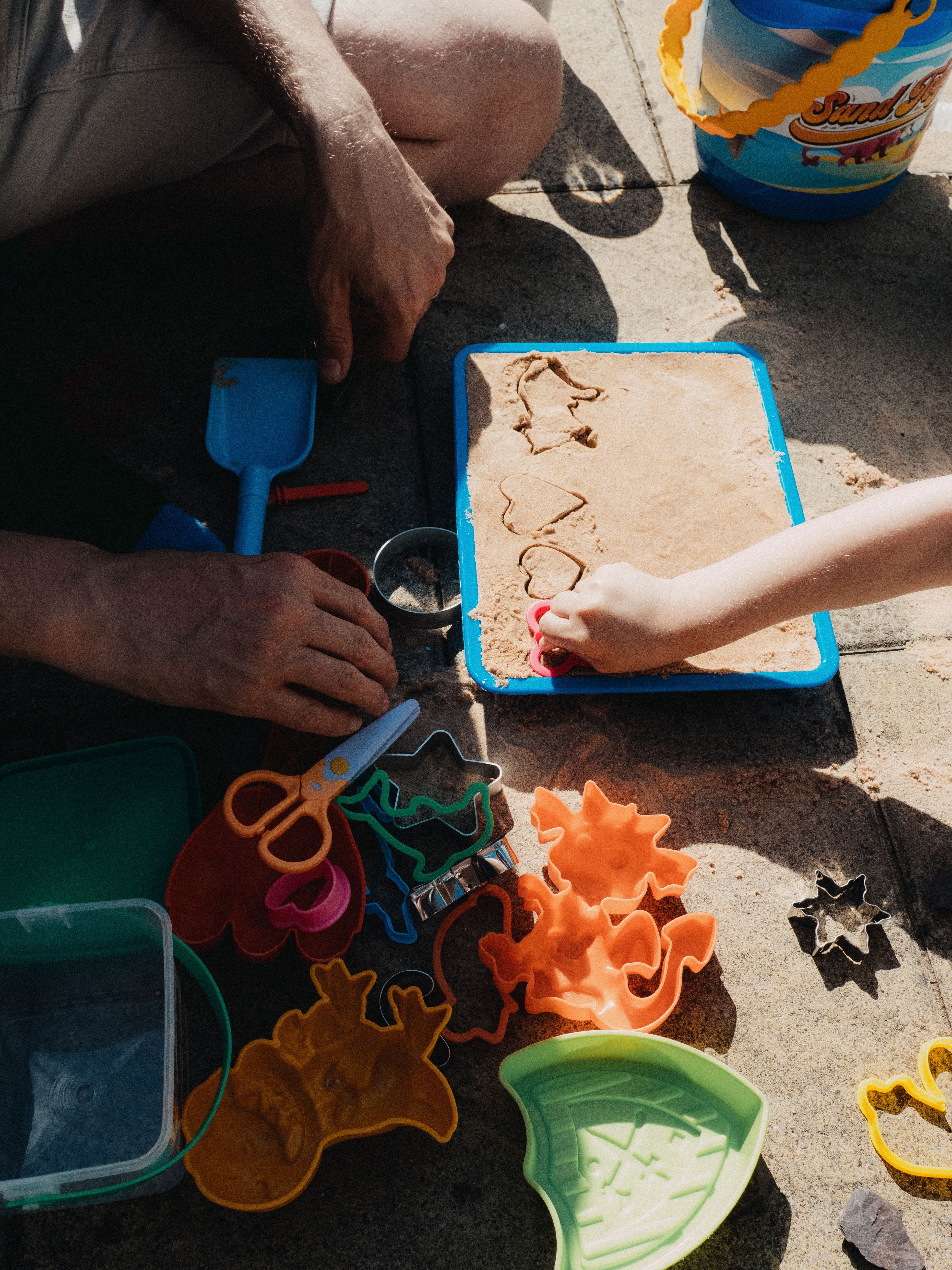 Father and child enjoying playtime with colorful sand toys on a sunny day outdoors.