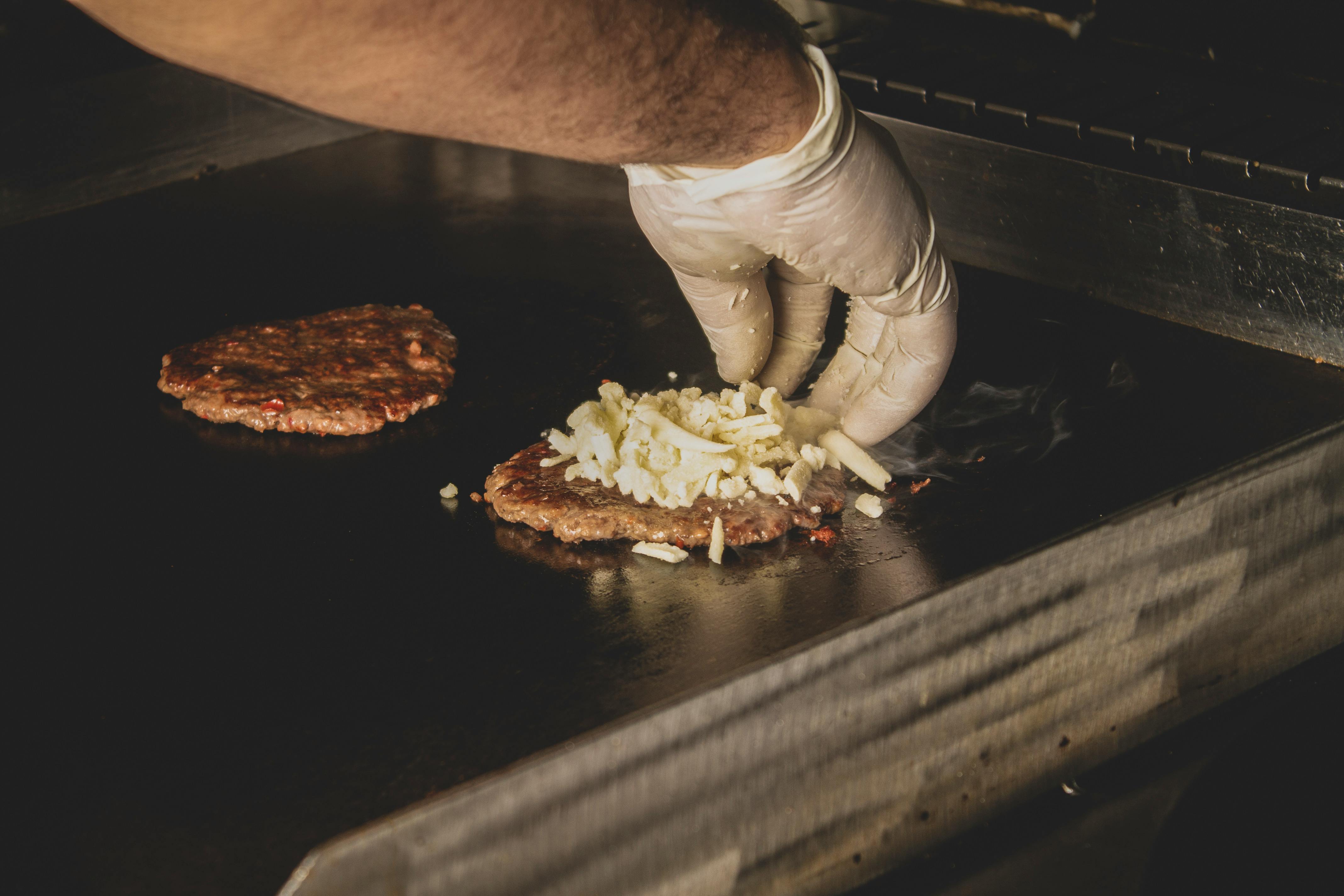 A chef hand in glove adding cheese to burger patties on a grill inside a commercial kitchen.
