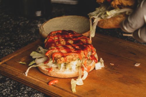 Free Close-up of a chicken burger being prepared with fresh lettuce and drizzled sauce on a wooden board. Stock Photo