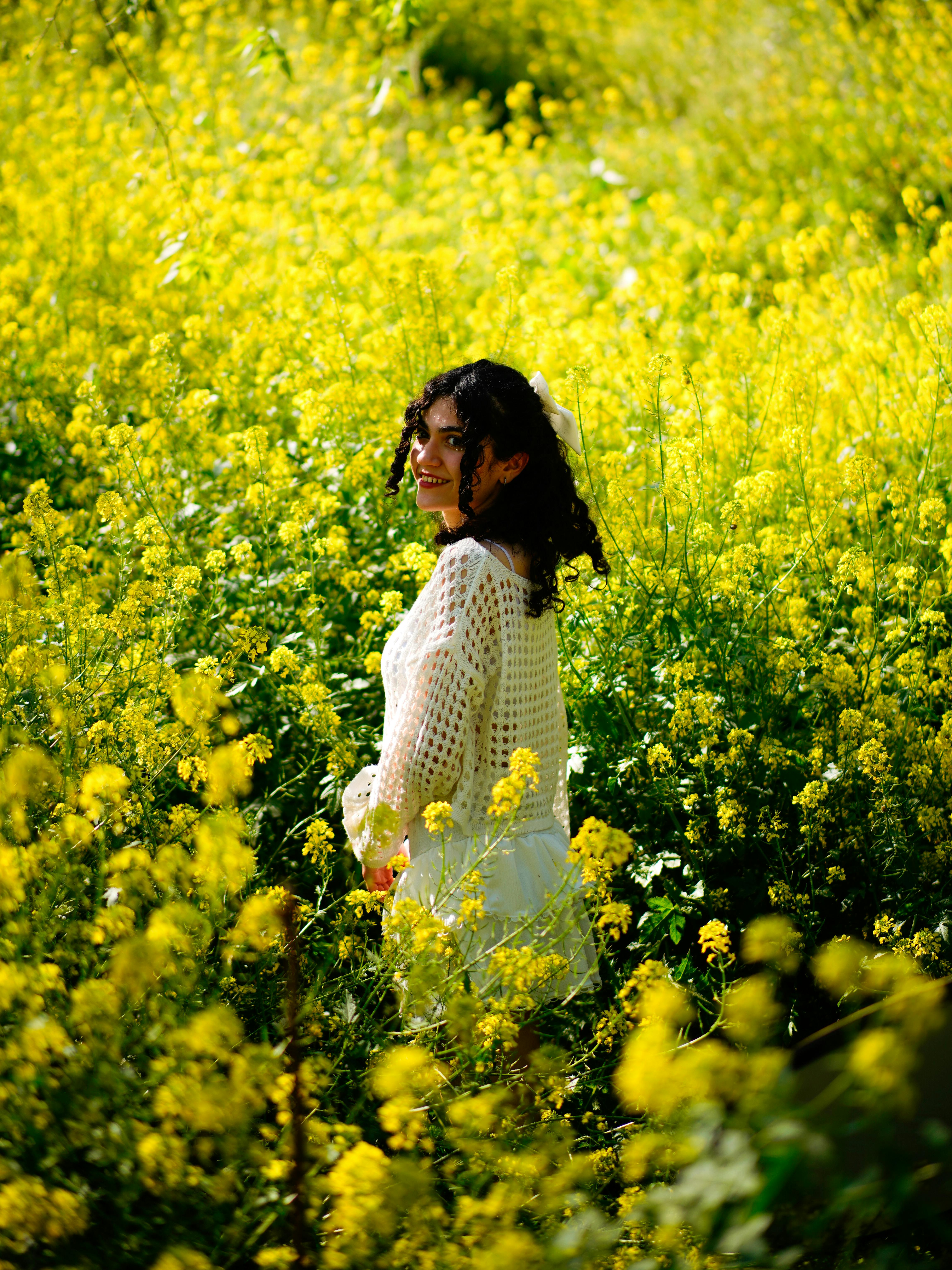 A woman smiling amidst a vibrant field of yellow flowers in a sunny outdoor setting.