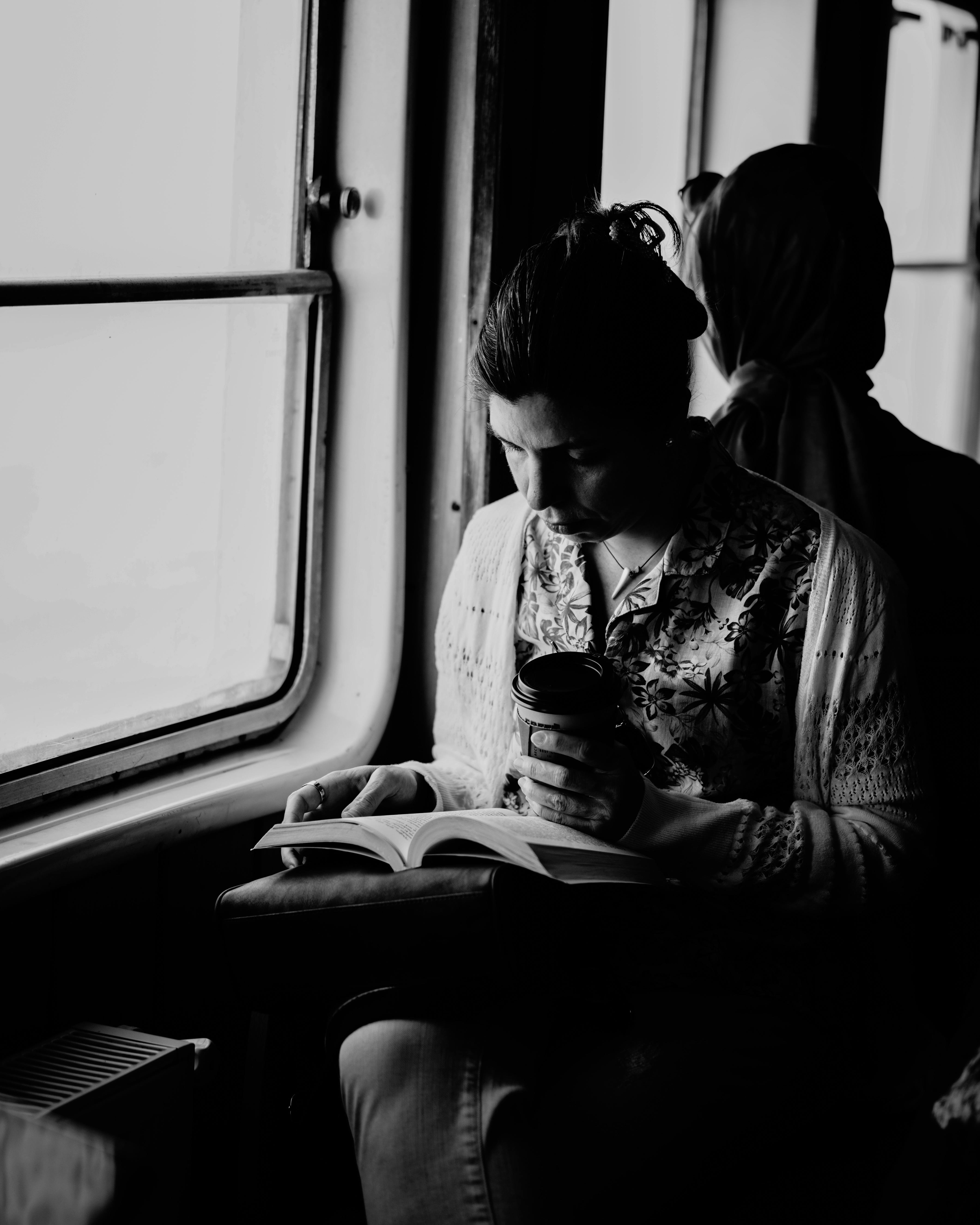 A woman reads a book with a coffee cup on a train in black and white.