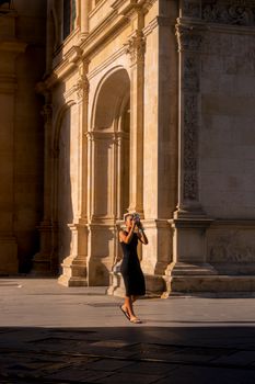 A woman in a black dress photographs historic architecture in warm sunlight.