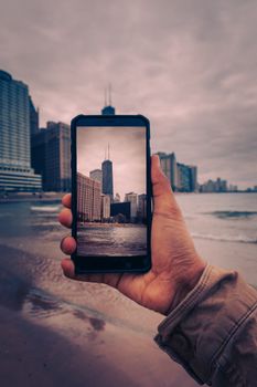 A hand holds a smartphone capturing a picturesque urban skyline along a beach.