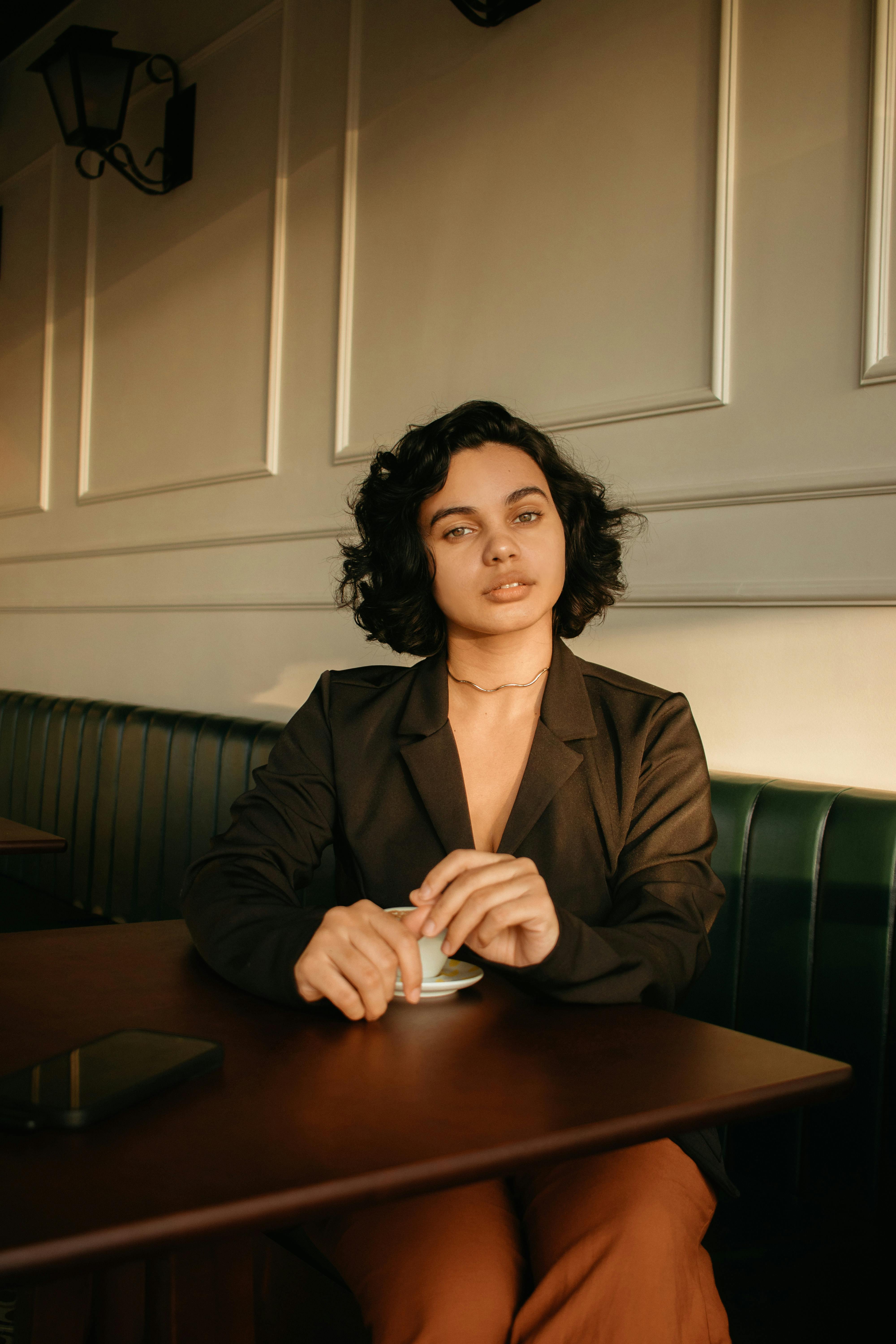 Woman in brown jacket sitting indoors at a cafe table with soft evening light.