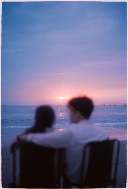 A couple sits on the beach at Vung Tau, Vietnam, embracing as the sun sets over the ocean.